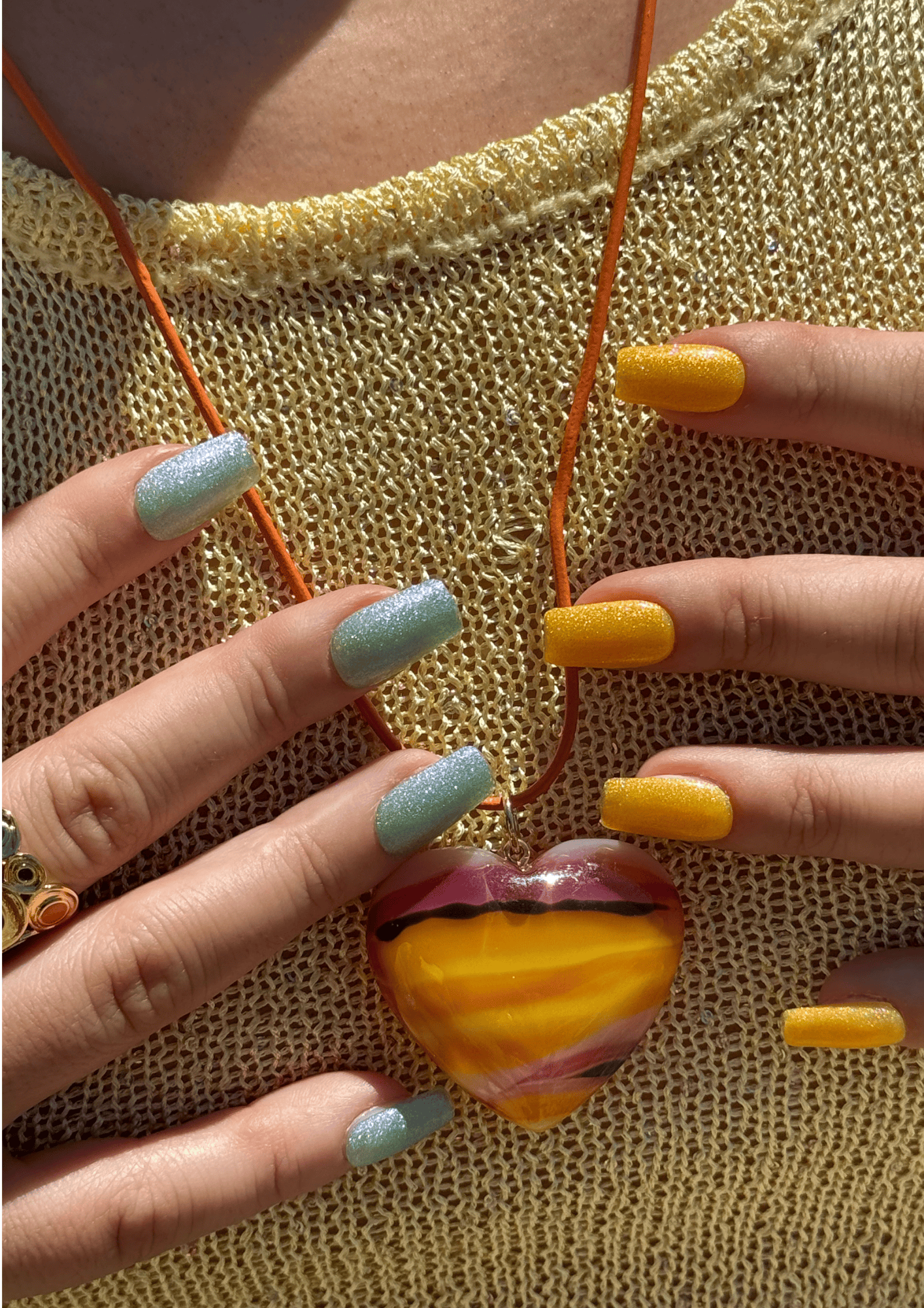 Two hands with mismatched glittery nails—light blue and golden yellow—holding a heart-shaped glass pendant with swirling orange and purple tones on an orange cord, against a gold knit top.