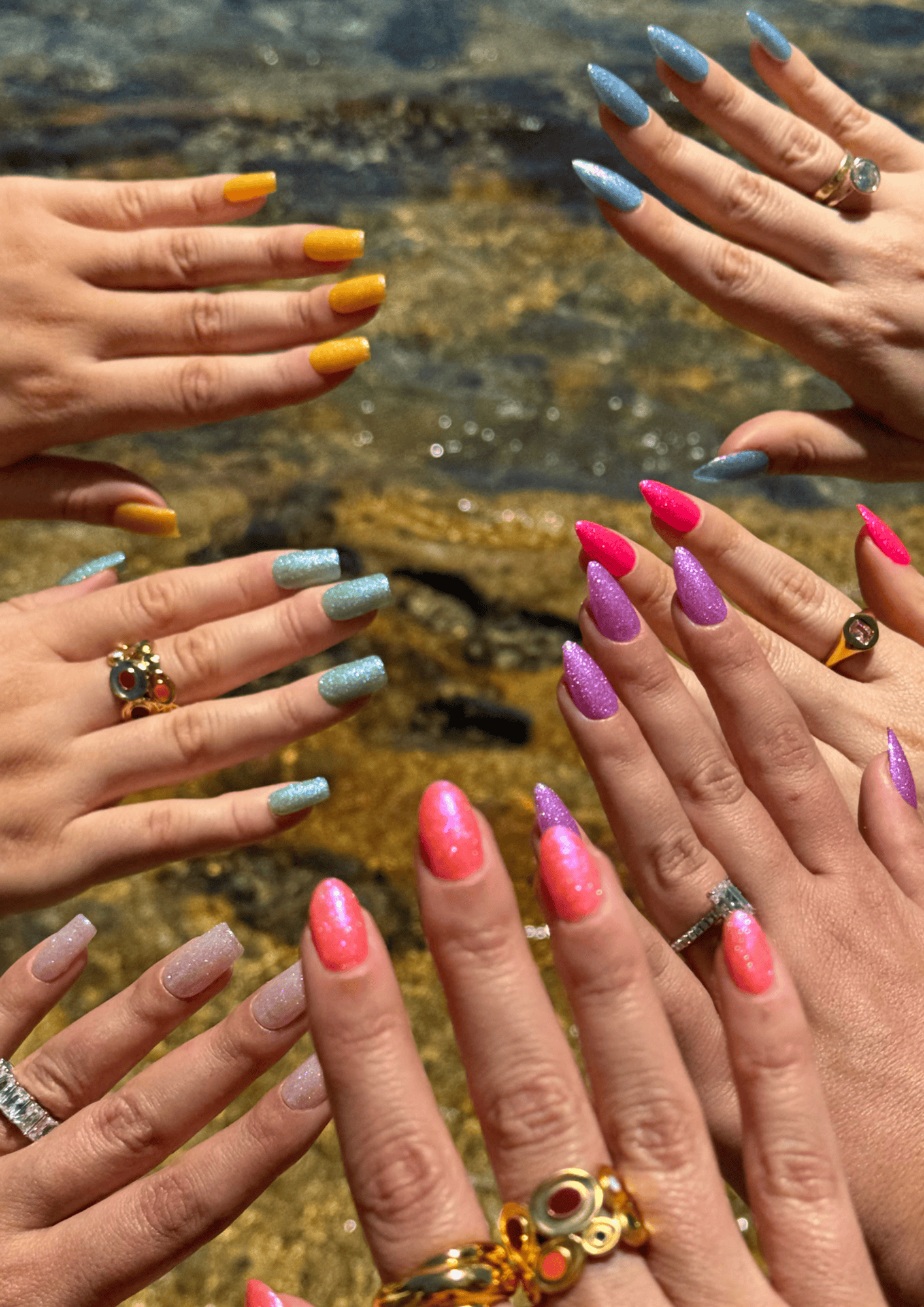 Six hands arranged in a circle displaying colorful glitter manicures in blue, orange, teal, hot pink, purple, and coral, with various rings on fingers, over a marble surface.