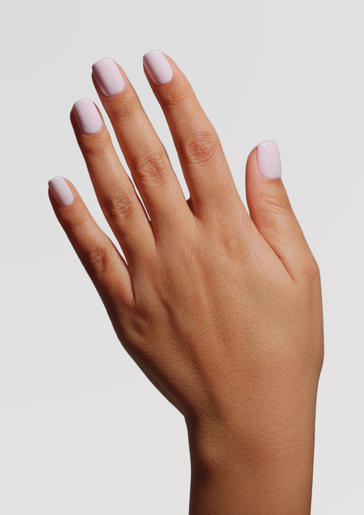 A hand with short, rounded nails coated in a glossy pale lavender-pink polish, raised against a light gray background.