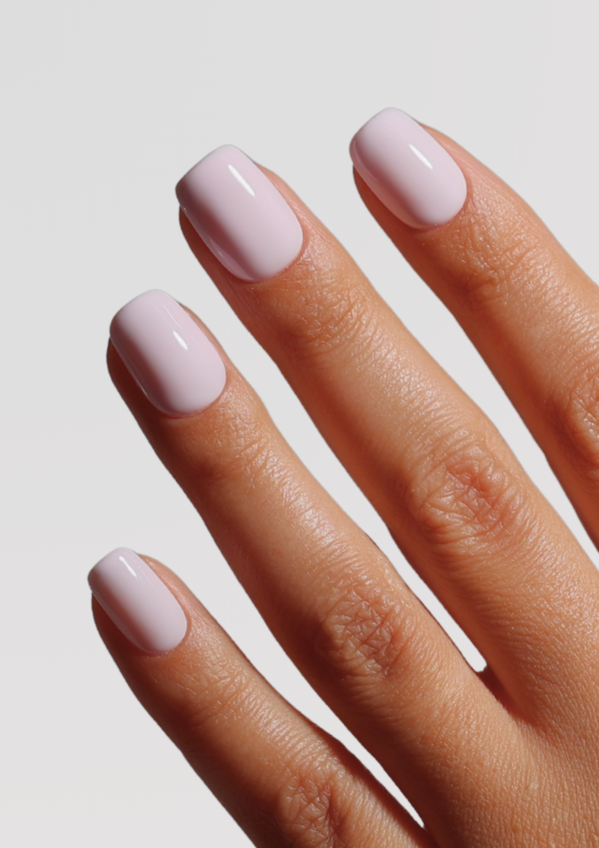 Close-up of a hand with four short, square-shaped nails coated in glossy pale lavender-pink polish against a light gray background.