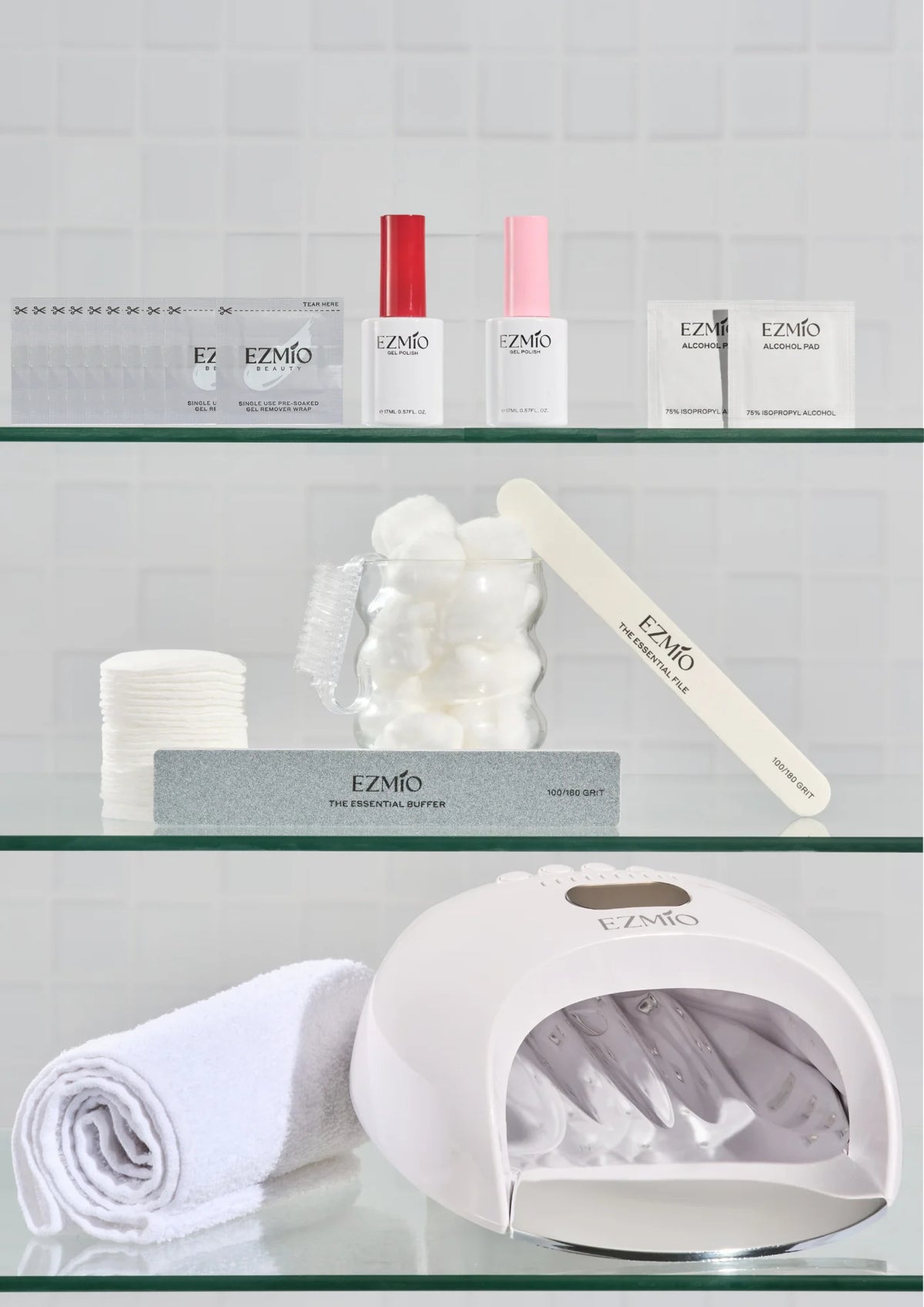 Three glass shelves displaying EZMio Gel Nail Kits nail products: gel polish bottles, gel remover wraps, alcohol pads, cotton pads, nail file, buffer block, a bear-shaped jar with cotton balls, and a white UV/LED nail lamp with a gloved hand inside.