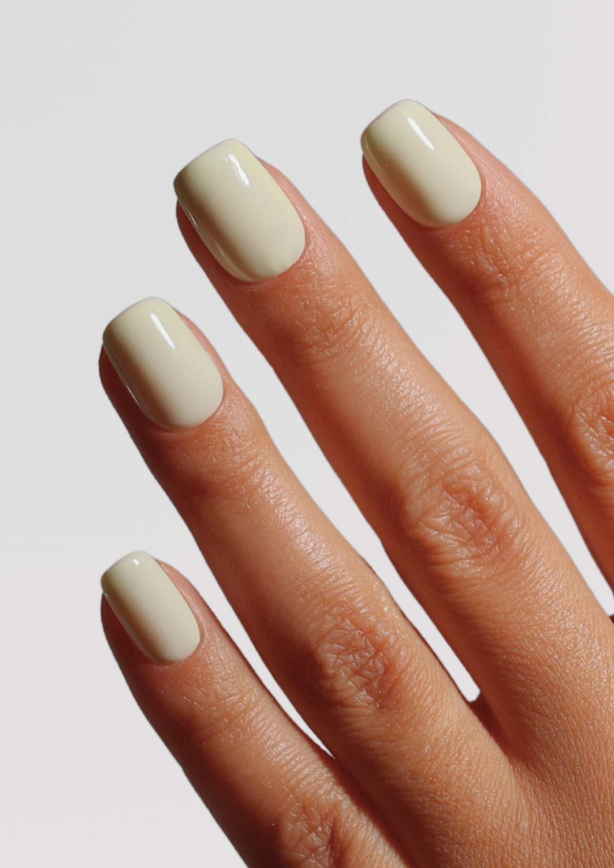 Close-up of a hand with four short, square-shaped nails coated in a glossy, creamy off-white nail polish against a light gray background.