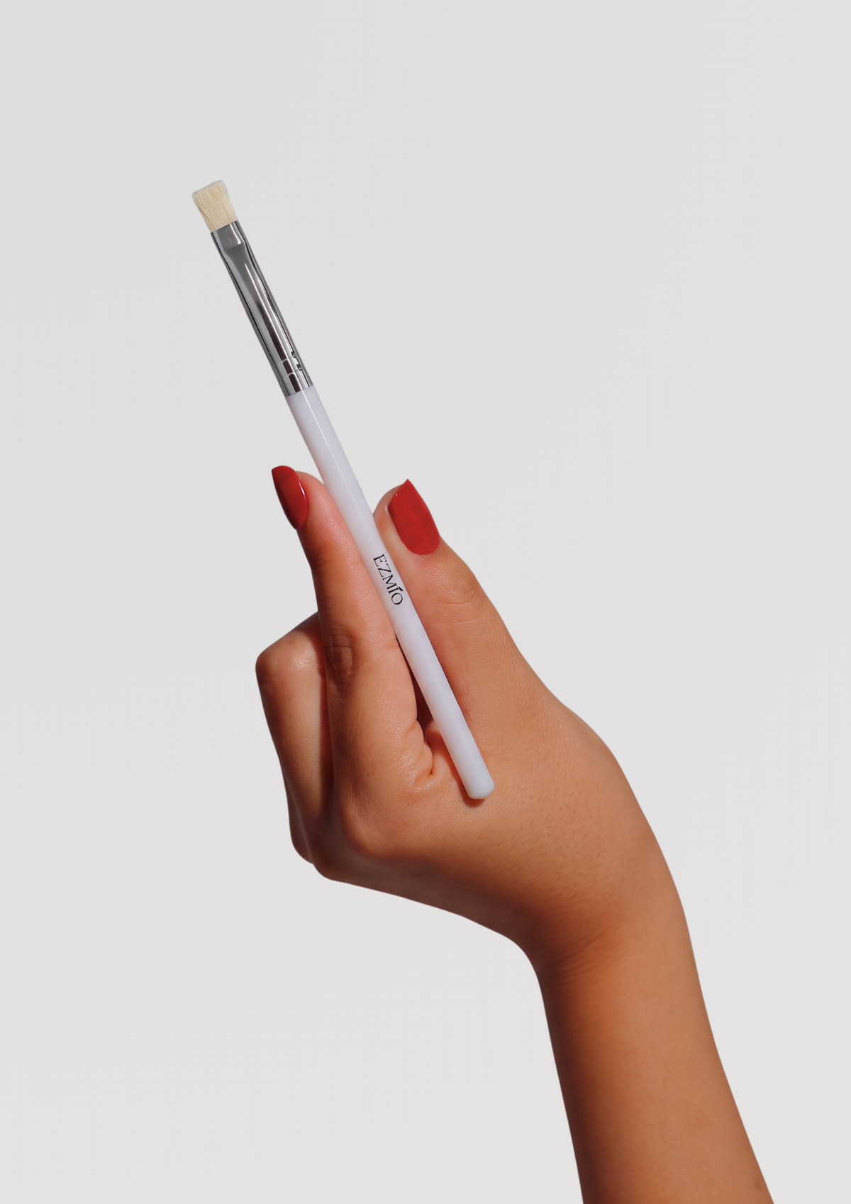 A hand with red-painted nails holds a slim makeup brush with a white handle, silver ferrule, and flat natural bristle tip, against a light gray background.