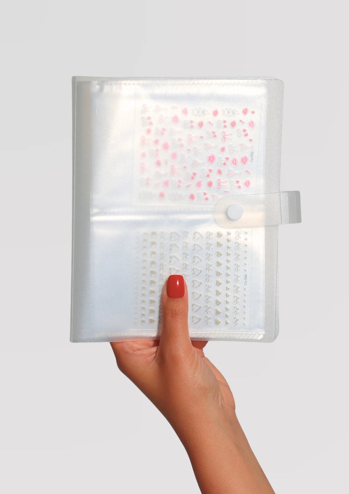 A hand with red nail polish holds a clear iridescent binder containing sheets of small pink and white/gold nail art stickers, secured with a snap closure.