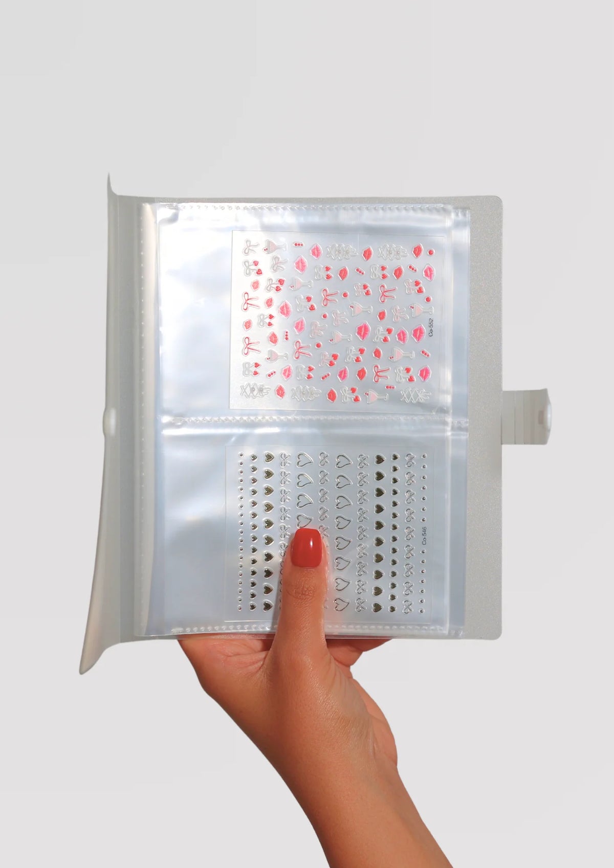 A hand with red nails holds a clear plastic organizer binder containing two sheets of nail art stickers: pink bows and lips on top, silver hearts and bows on the bottom.