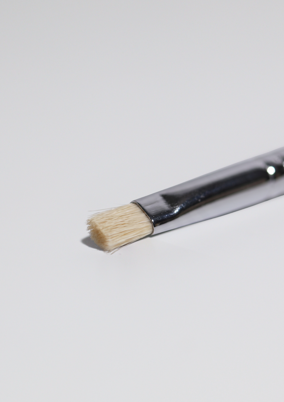 Small flat brush with cream-colored natural bristles and a shiny silver metallic handle, lying diagonally on a light gray surface.